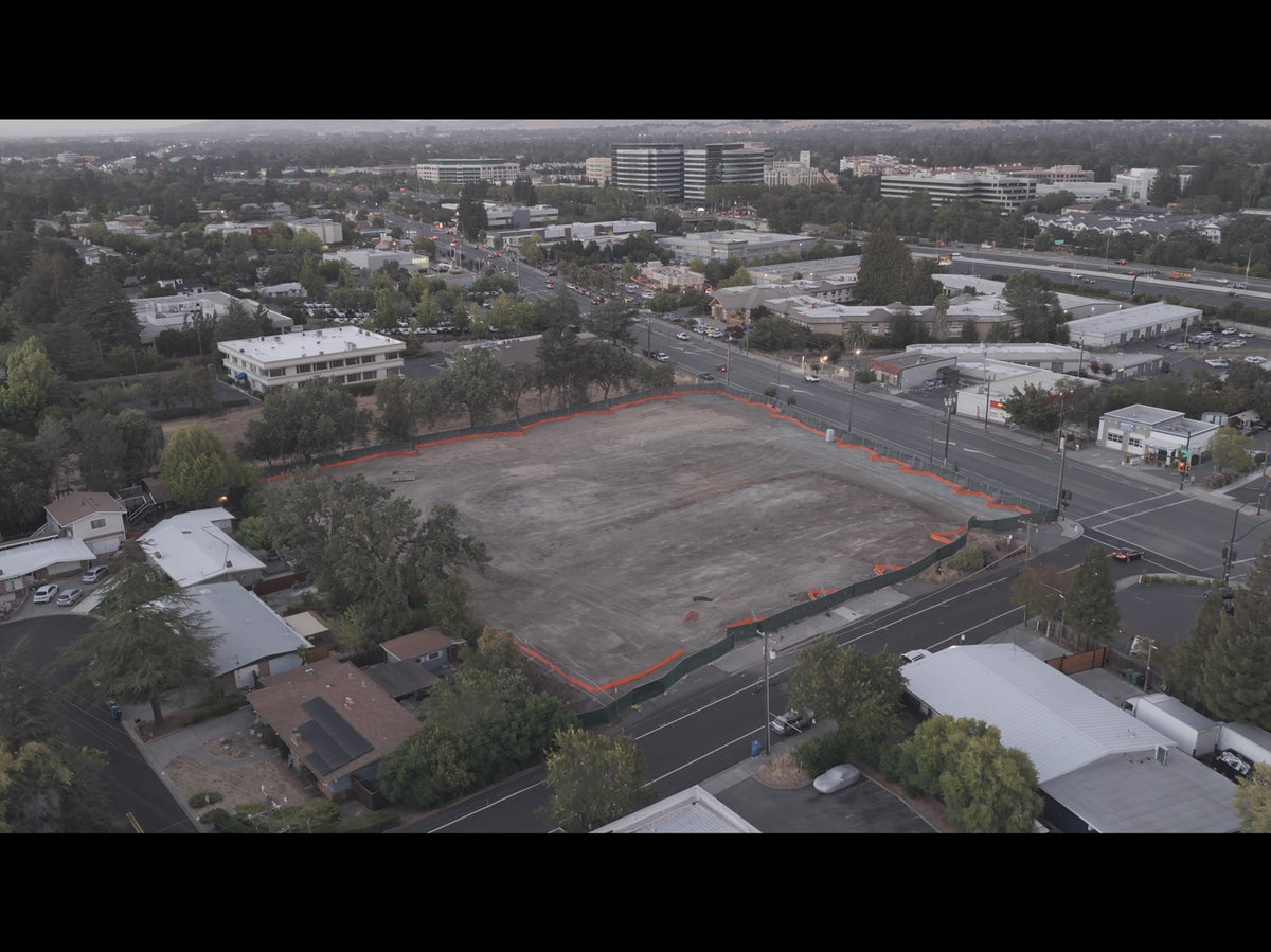 An aerial view of a large vacant lot outlined in orange tape, surrounded by roads, trees, and nearby commercial buildings.