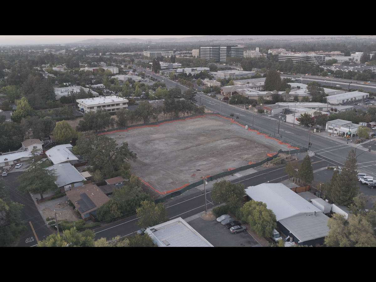 Aerial view of an empty rectangular lot outlined in orange construction fencing, surrounded by roads, trees, and commercial buildings.