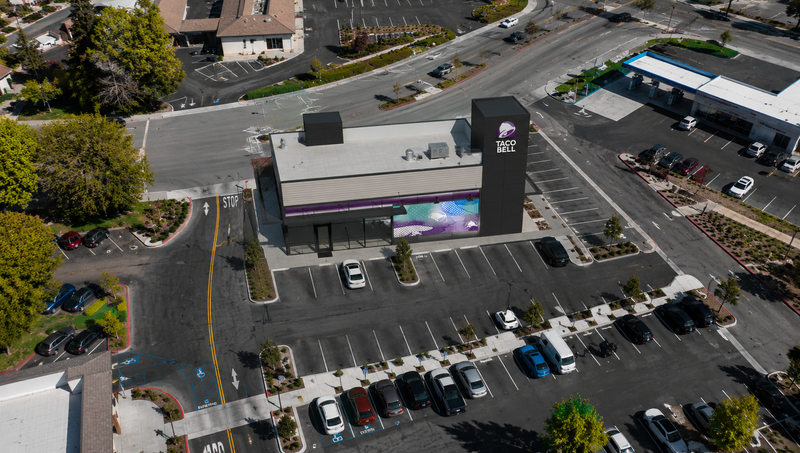 Aerial view of a 7-Eleven convenience store building with a large purple and blue mural on the side, surrounded by parking lots and nearby commercial buildings.