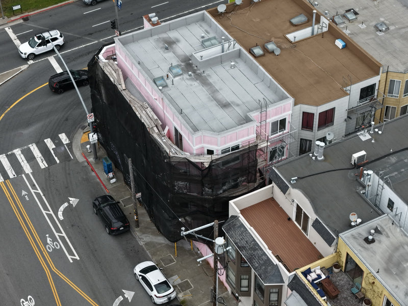 An aerial view of a pink building corner wrapped in black construction netting, situated at an intersection with streets, cars, and neighboring buildings visible.