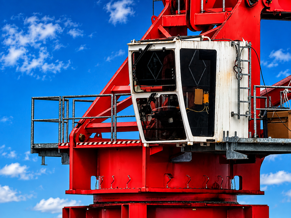 Close-up of a red industrial crane or gantry structure with a white operator cab and metal railings, photographed against a bright blue sky with scattered clouds.