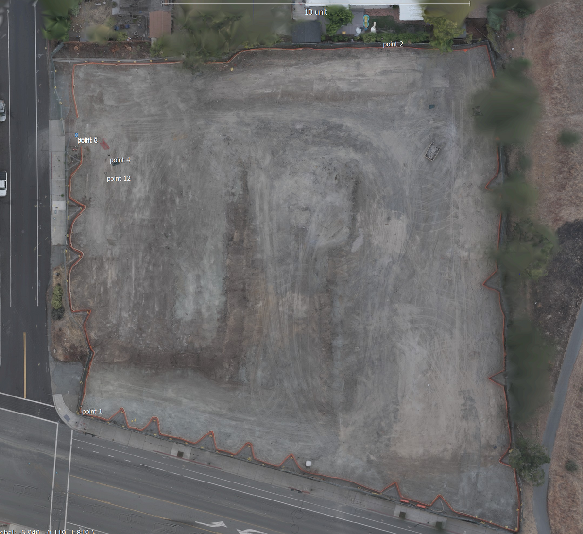 Aerial view of an undeveloped lot or construction site with dirt ground, orange safety fencing along the perimeter, adjacent roads, and some trees. Small text markers are visible on the left side of the lot.