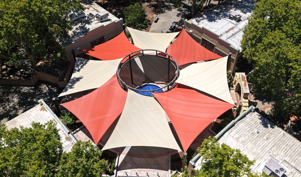 An aerial view of a large star-shaped canopy structure with alternating red and beige fabric panels surrounding a central open ring, installed over a building with surrounding trees and rooftops visible.