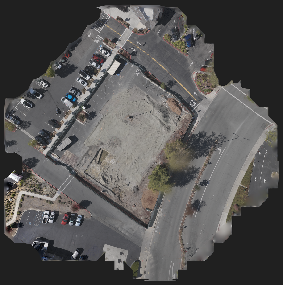 Aerial view of a vacant lot or construction site with a dirt patch in the center, surrounded by paved parking areas, roads, and landscaping, isolated on a black background.