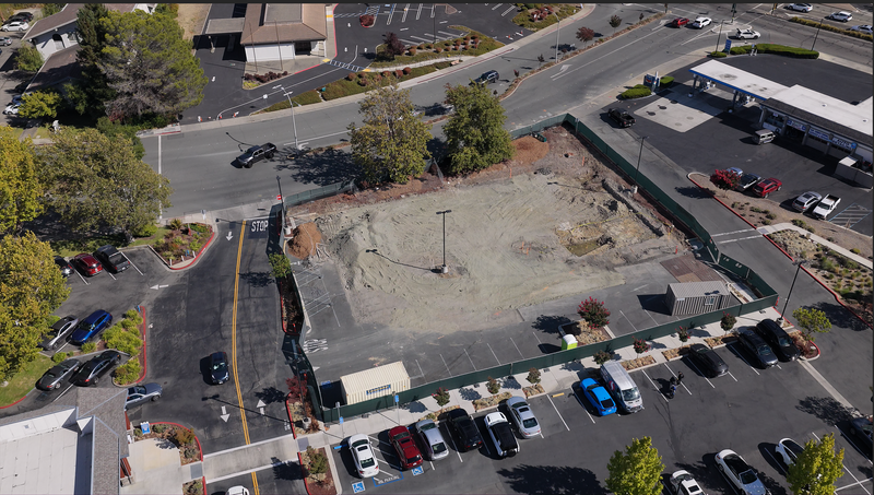 Aerial view of a vacant, fenced lot with a large patch of bare dirt in the center, surrounded by a parking lot, roads, trees, and nearby commercial buildings.