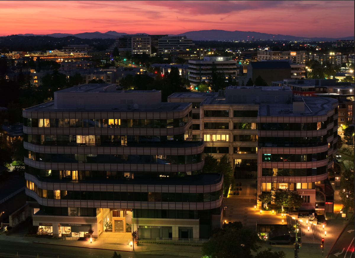 An aerial view of a modern multi-story office building complex at dusk, with glowing yellow windows, surrounding city lights, and mountains under a pink and orange sunset sky.