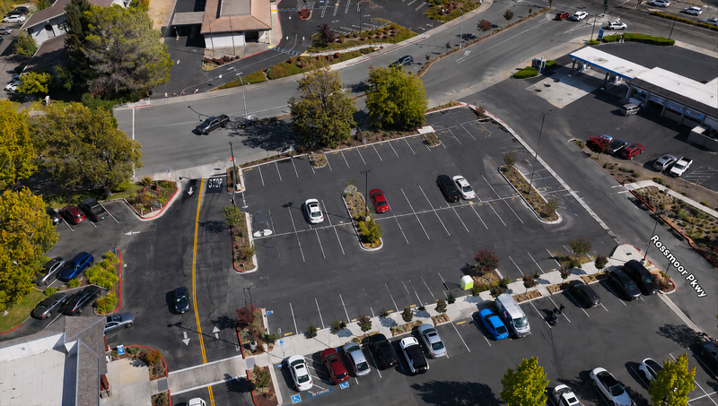 Aerial view of a commercial parking lot with multiple parked cars, landscaped islands, roads, and adjacent buildings.